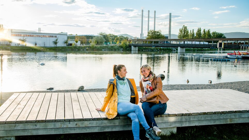 Two women relax by the water in Graz, surrounded by nature. | © Graz Tourismus
