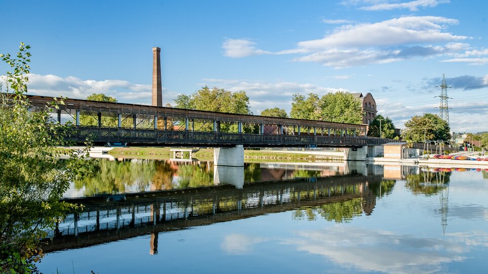 A bridge over the calm Mur, surrounded by trees and historic buildings. | © Graz Tourismus