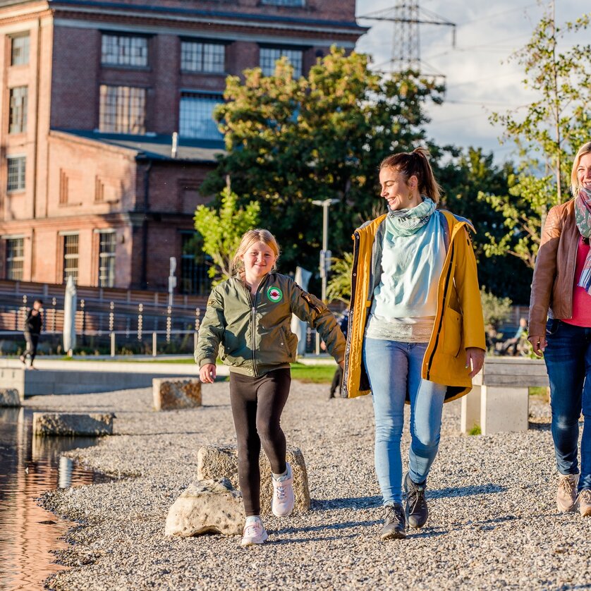 Two women and a girl walk along the banks of the Mur in Graz. The soap factory can be seen in the background. | © Graz Tourismus