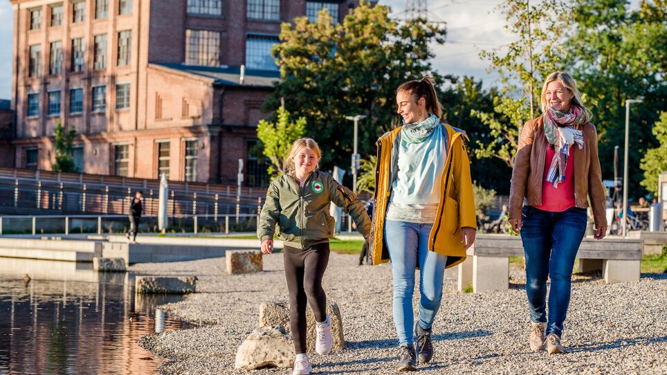 Two women and a girl walk along the banks of the Mur in Graz. The soap factory can be seen in the background. | © Graz Tourismus