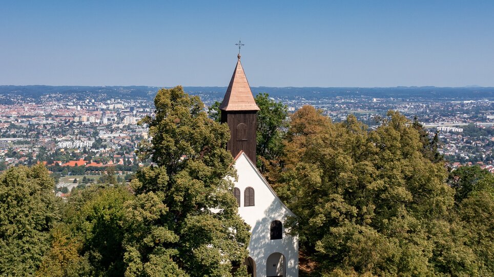 Die Kirche St. Johann und Paul -umgeben von Bäumen mit Sicht auf Graz. | © Graz Tourismus