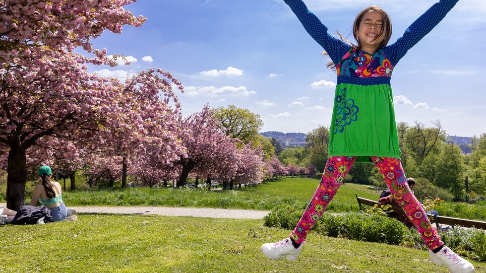 A girl joyfully jumps in the park among cherry blossom trees.
