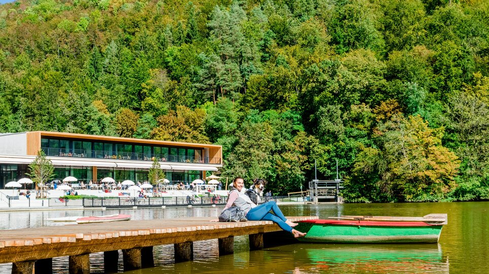 Frau sitzt auf einem Steg am Thalersee, umgeben von Bäumen und einem Café im Hintergrund. | © Graz Tourismus