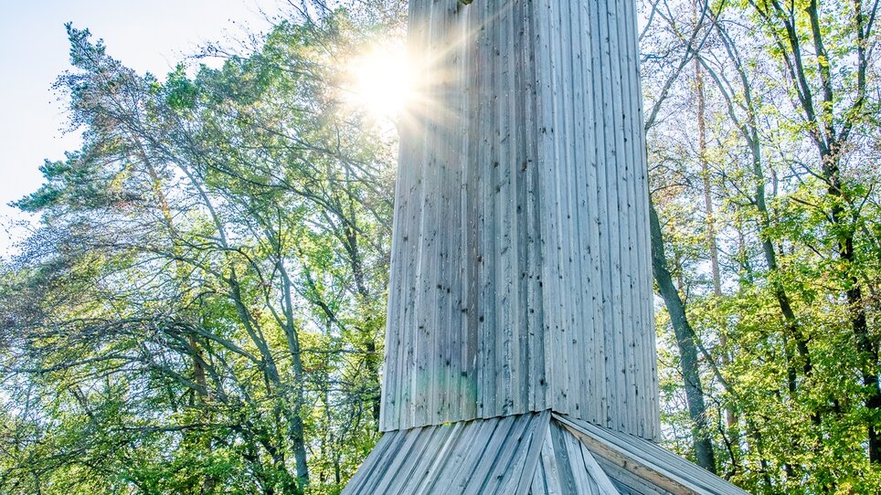 Zwei Frauen sitzen an einer Holzbank neben dem Wetterturm Schaftalberg - einem hohen Holzbau mit Kreuz, umgeben von Bäumen. | © Graz Tourismus