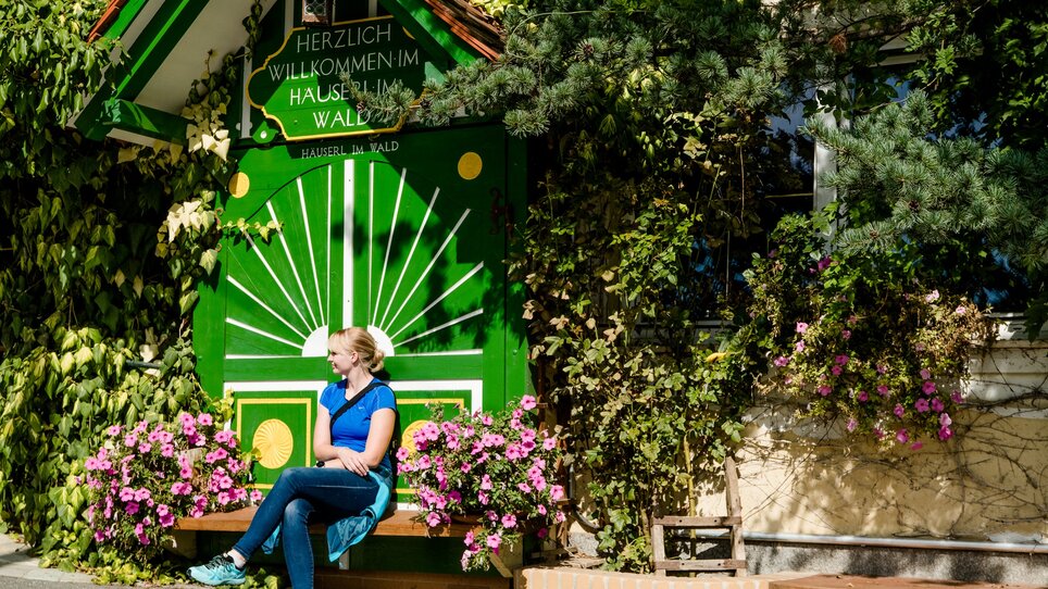 Frau sitzt vor einem grünen Schild des Restaurants "Häuserl im Wald" - umgeben von blühenden Pflanzen. | © Graz Tourismus