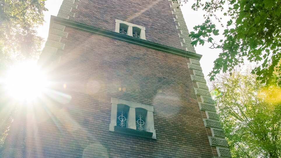 Blick auf die Hilmwarte - einen hohen Turm umgeben von Bäumen mit Sonnenschein im Hintergrund. | © Graz Tourismus