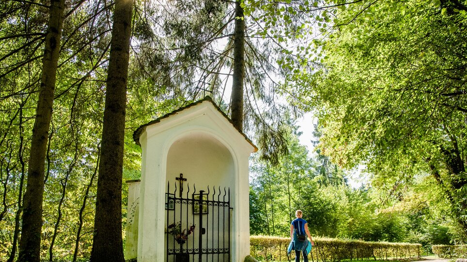 Person läuft durch einen sonnigen Wald - vorbei an einer weißen Kapelle. | © Graz Tourismus