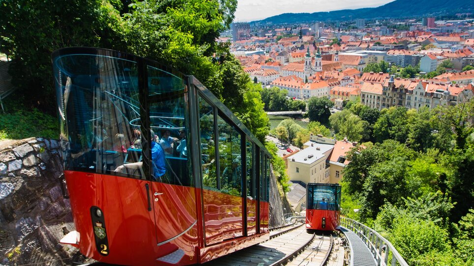 Zwei rot-schwarze Bahnen fahren den Schlossberg in Graz hinunter, die Stadt und die Mur im Hintergrund. | © Graztourismus - Harry Schiffer