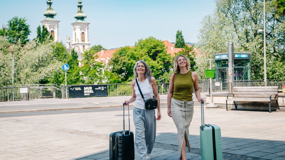Zwei Frauen gehen lächelnd mit Koffern durch Graz, bei sonnigem Wetter. | © Graz Tourismus - Mias Photoart