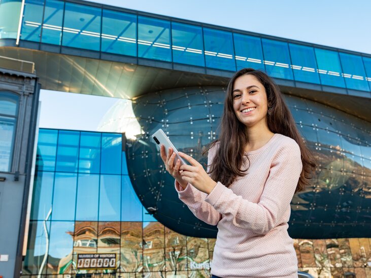 A young woman smiles holding her smartphone in front of a reflective building in Graz. | © Graz Tourismus - Harry Schiffer