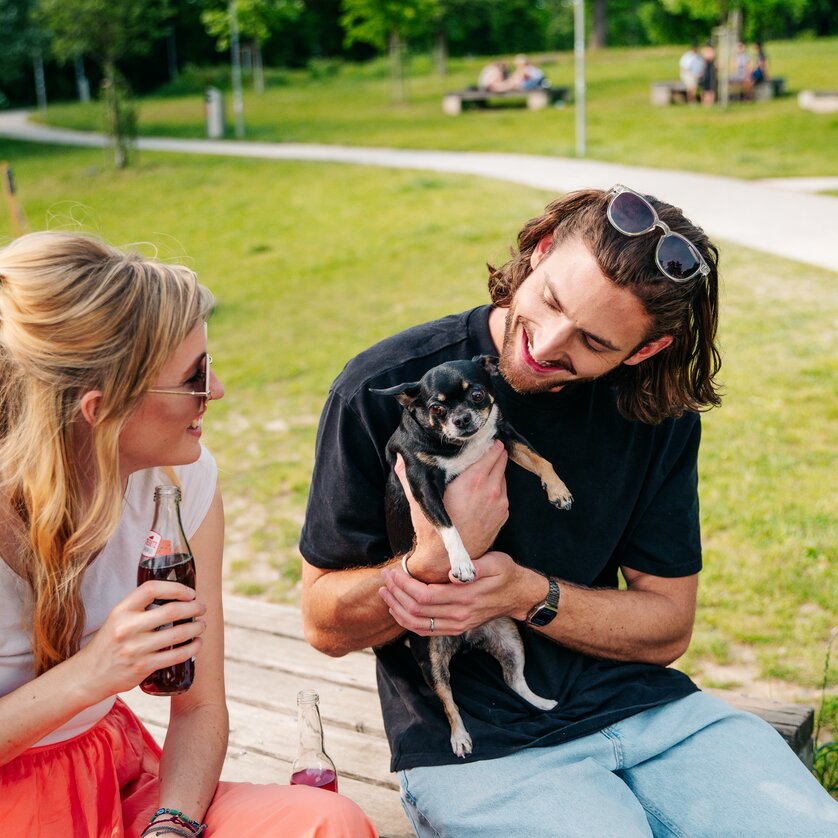 Pärchen im Augarten mit Hund und Getränken, entspannte Atmosphäre. | © Graz Tourismus - Mias Photoart