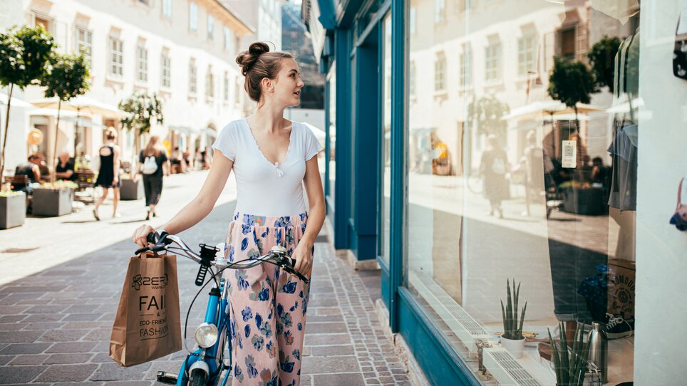 A young woman in floral pants stands with a bicycle in front of a shop window in Graz. | © Graz Tourismus