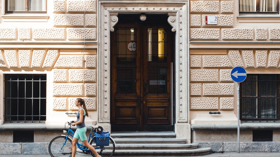 Eine Frau schiebt ein Fahrrad an einem historischen Gebäude in Graz vorbei. | © Graz Tourismus