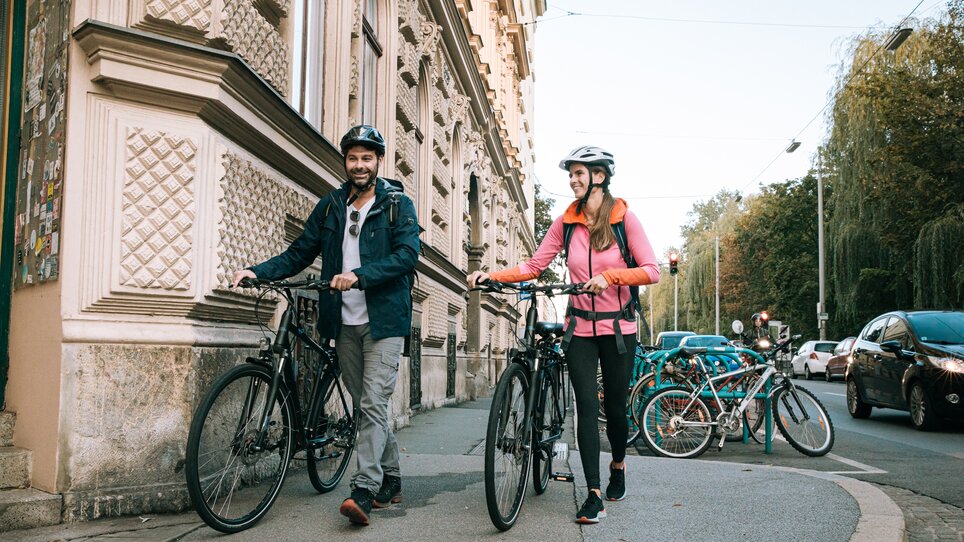 Zwei Radfahrer schieben ihre Fahrräder in Graz, umgeben von Gebäuden. | © Graz Tourismus