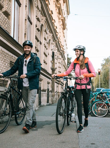 Two cyclists walk their bikes in Graz, surrounded by buildings. | © Graz Tourismus