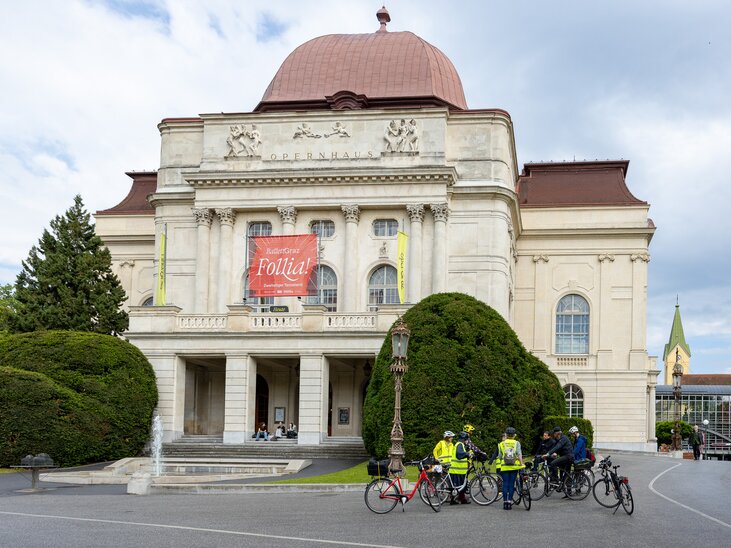 Das Opernhaus in Graz mit Radfahrern davor und einem großen Plakat. | © Graz Tourismus - Harry Schiffer