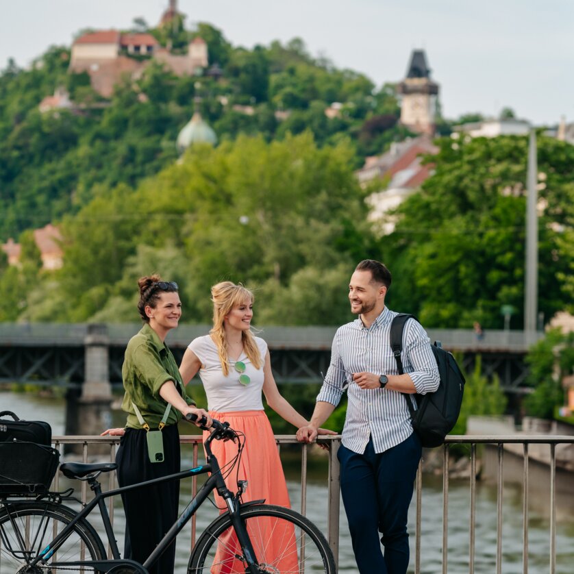 Cycling I Sport & Leisure in Graz | © Graz Tourismus - Mias Photoart Three people with bicycles in front of the Mur and the Graz Clock Tower. | © Graz Tourismus - Mias Photoart