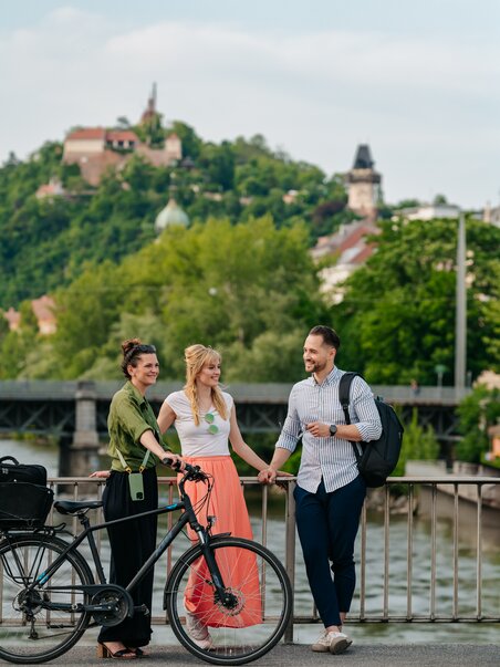 Drei Menschen mit Fahrrädern vor der Mur und dem Grazer Uhrturm. | © Graz Tourismus - Mias Photoart