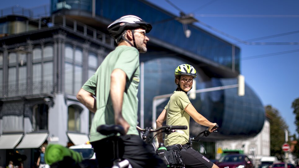 Two cyclists smiling in Graz. Modern architecture in the background. | © Graz Tourismus - Tom Lamm