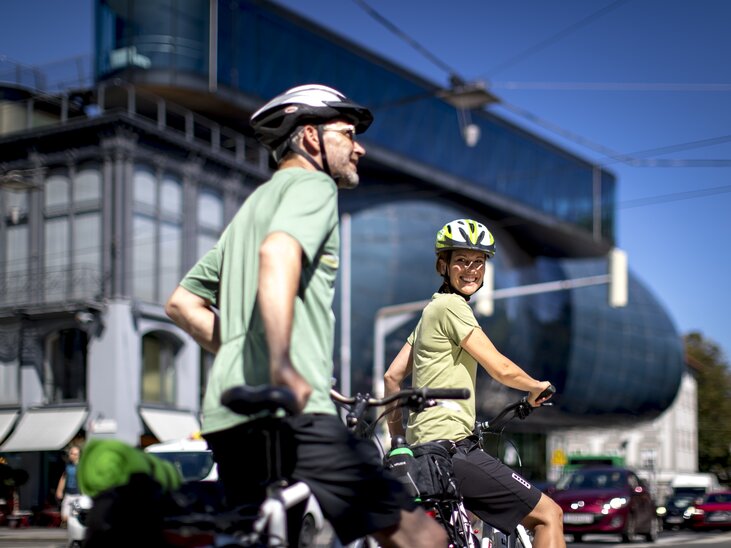 Two cyclists smiling in Graz. Modern architecture in the background. | © Graz Tourismus - Tom Lamm