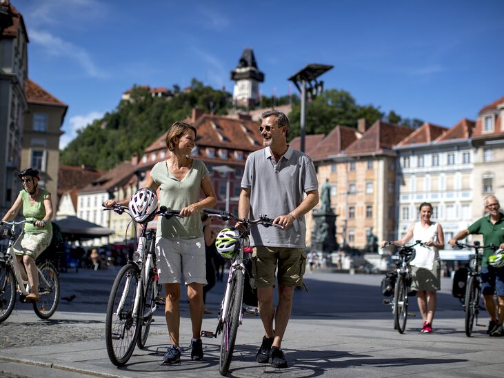 Paar und weitere Radfahrer in Graz, mit dem Grazer Uhrturm im Hintergrund. | © Graz Tourismus - Tom Lamm