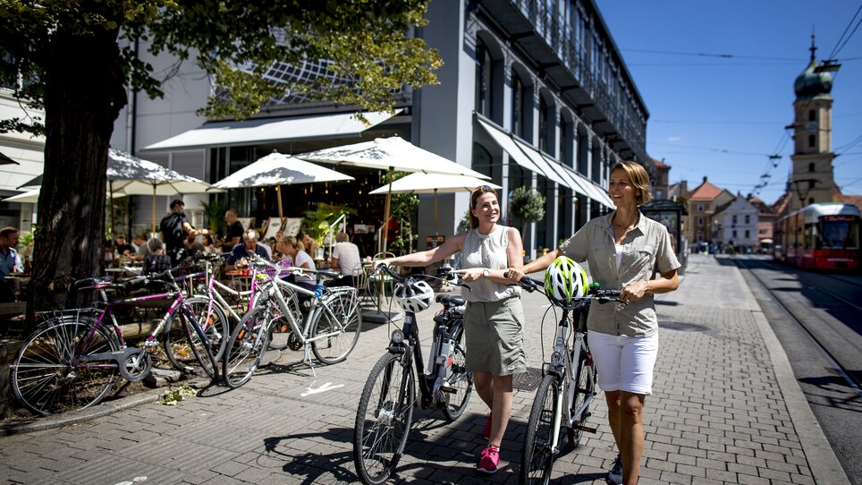 Zwei Frauen schieben Fahrräder neben dem Kunsthaus in Graz.