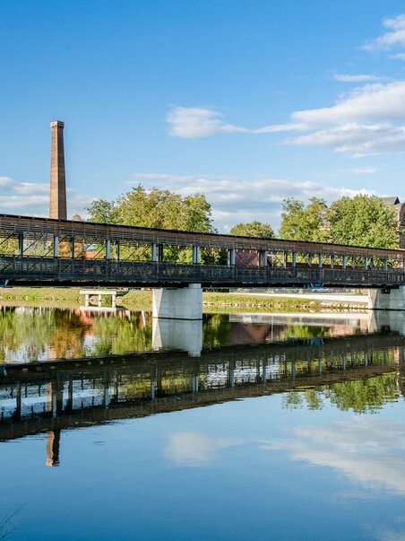 Eine Brücke über die Mur, idyllische Landschaft mit reflektierendem Wasser. | © Graz Tourismus - Mias Photoart