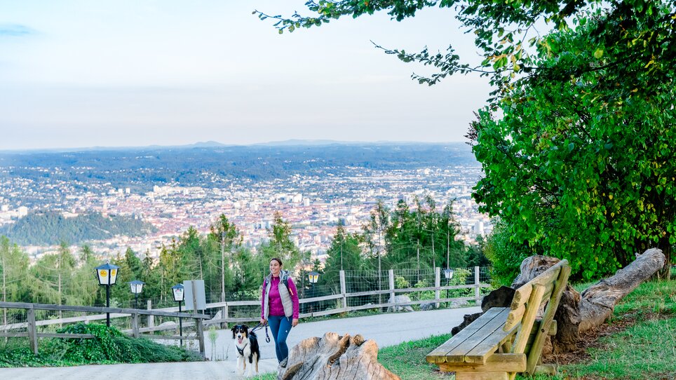 A woman walks with her dog on a path overlooking Graz. | © Graz Tourismus - Mias Photoart