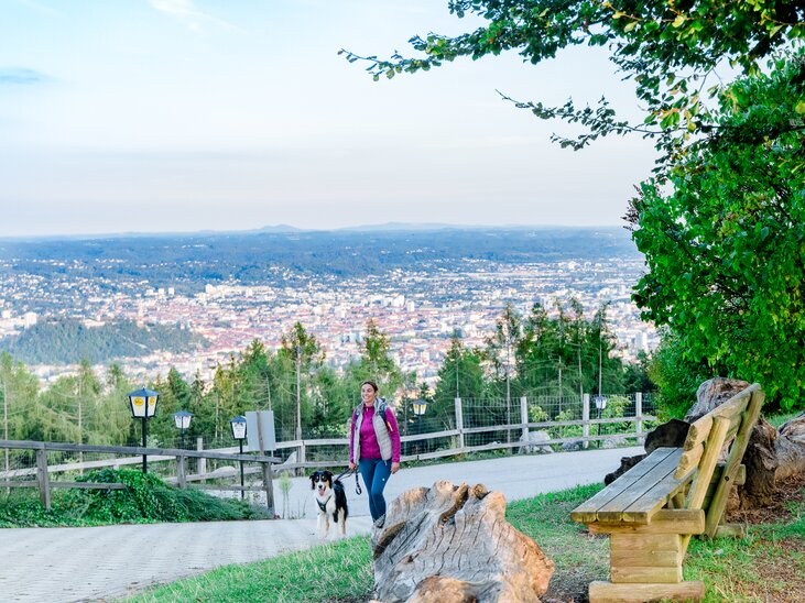 Eine Frau geht mit ihrem Hund auf einem Weg mit Aussicht über Graz. | © Graz Tourismus - Mias Photoart