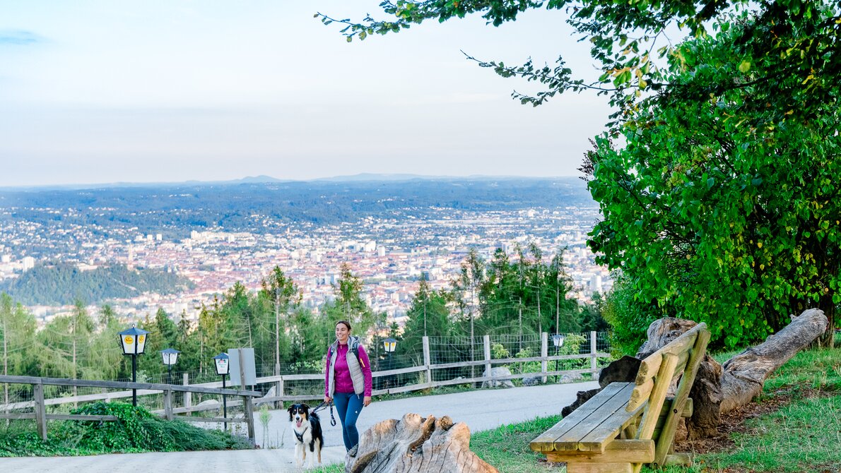 A woman walks with her dog on a path overlooking Graz. | © Graz Tourismus - Mias Photoart