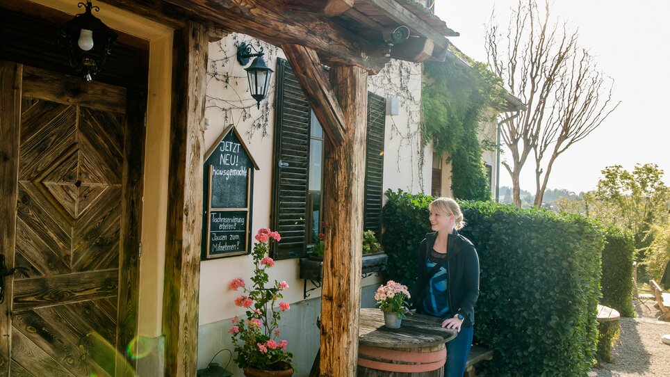 A woman stands in front of a rustic building with flowers. | © Graz Tourismus - Mias Photoart