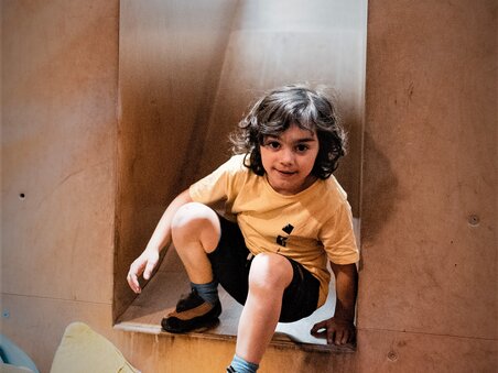 Ein Kind krabbelt durch einen rechteckigen Ausschnitt in der Wand, umgeben von bunten Kissen. | © Clemens Bender