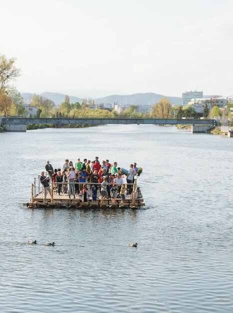 Gruppo di persone su una zattera sul fiume Mur a Graz. Atmosfera rilassata accanto all'acqua. | © LippZahnschirm