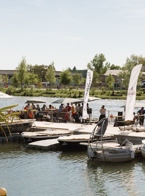 A busy jetty on the banks of the Mur in Graz with lots of people and boats. | © Die Flößerei