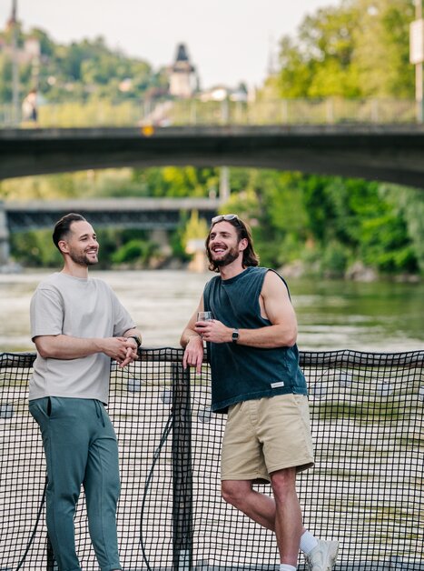 Zwei Männer auf einem Floß auf der Mur in Graz, umgeben von Natur und einer Brücke. Im Hintergrund sieht man den Grazer Uhrturm. | © Graz Tourismus - Mias Photoart