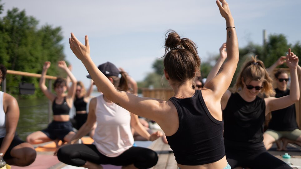 Un gruppo di persone partecipa a un corso di yoga su una zattera sul fiume Mur a Graz, organizzato da “Der Flößerei”. | © Robert Supnig