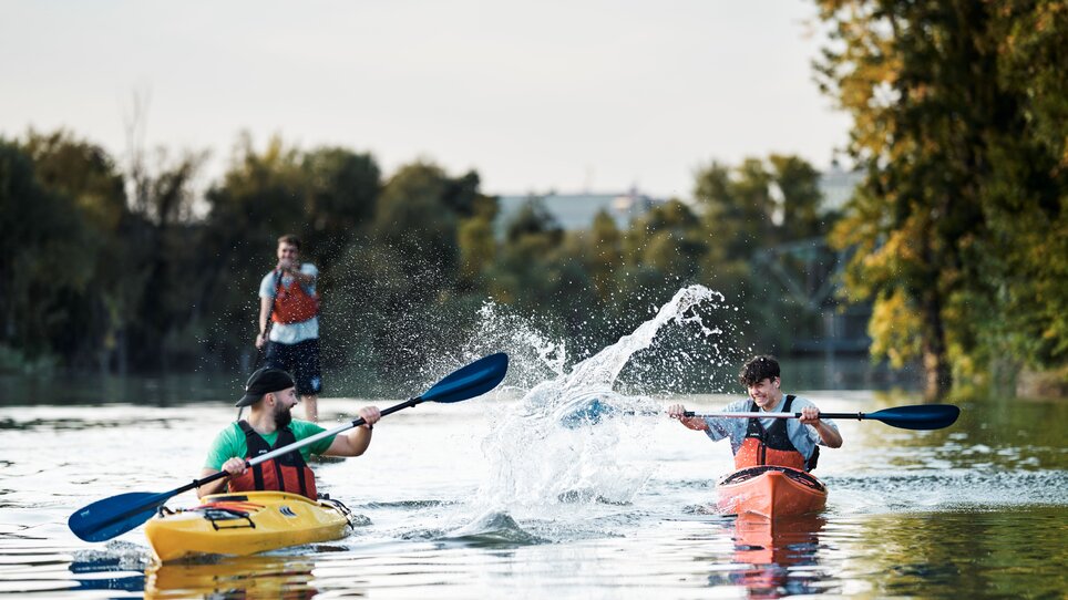 Zwei Kajakfahrer paddeln auf ruhigem Wasser, während ein Dritter auf einem SUP steht und zuschaut. | © Stadtstrand Graz - Stefan Leitner