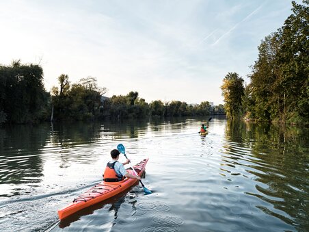 Two people in a kayak paddling on the calm Mur surrounded by trees. | © Stadtstrand Graz - Stefan Leitner