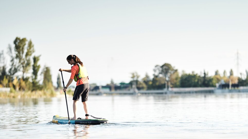 Eine junge Frau paddelt auf einem Stand-up-Paddleboard auf ruhig fließendem Wasser. | © Stadtstrand Graz - Stefan Leitner