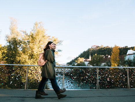 Ein Pärchen spaziert über eine Brücke in Graz mit Blick auf die Mur und den Grazer Uhrturm. | © Graz Tourismus
