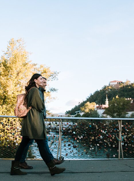 A woman and a man walk over a bridge in Graz, overlooking the Mur and the Graz Clock Tower. | © Graz Tourismus