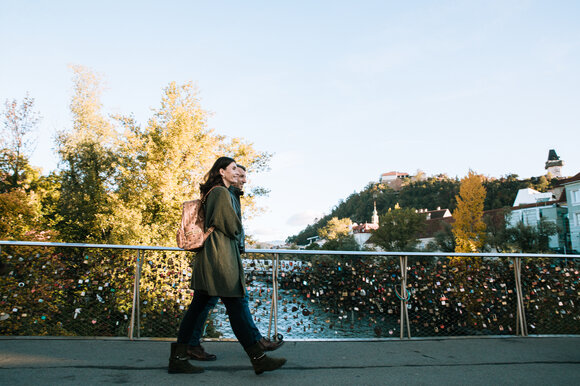 Ein Pärchen spaziert über eine Brücke in Graz mit Blick auf die Mur und den Grazer Uhrturm. | © Graz Tourismus