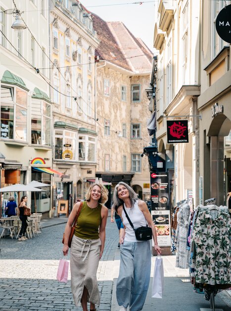 Due donne sorridenti mentre fanno shopping a Graz, con borse. | © Graz Tourismus - Mias Photoart
