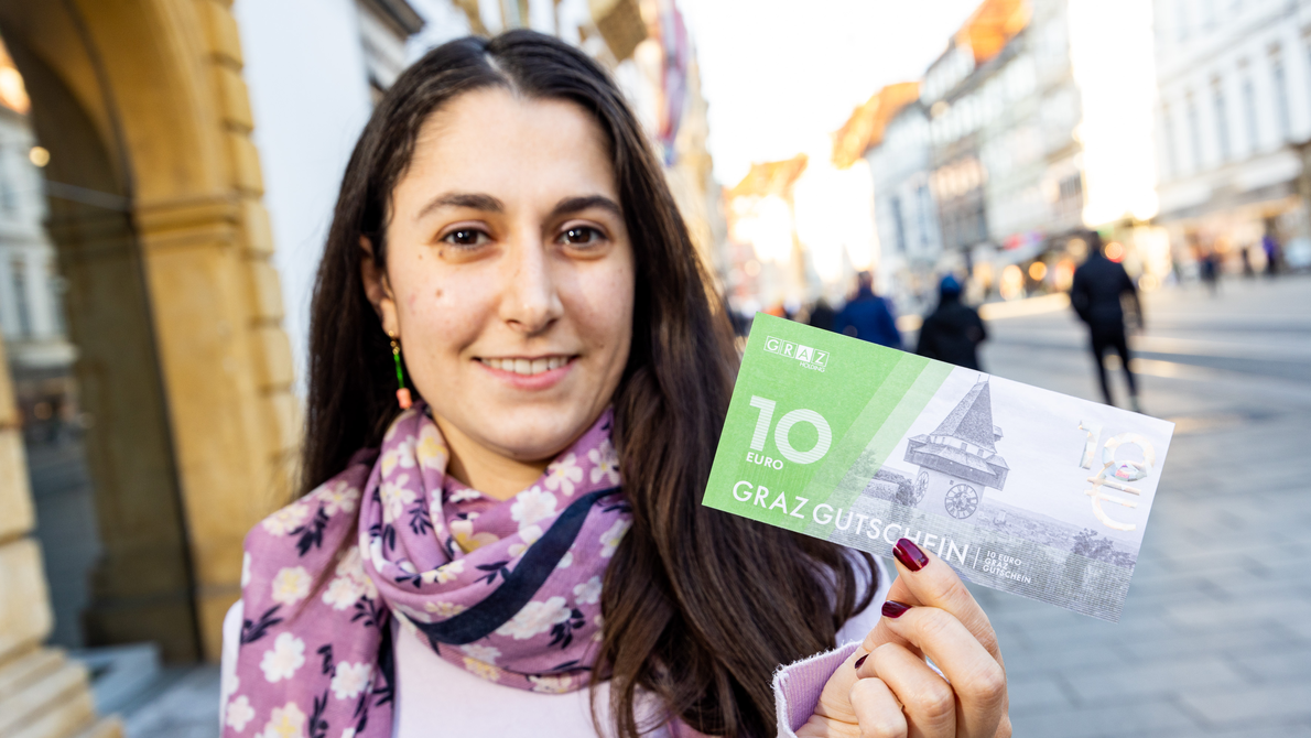 Eine Frau steht in der Grazer Herrengasse und hält einen 10 Euro Graz Gutschein in der Hand. | © Graz Tourismus - Harry Schiffer