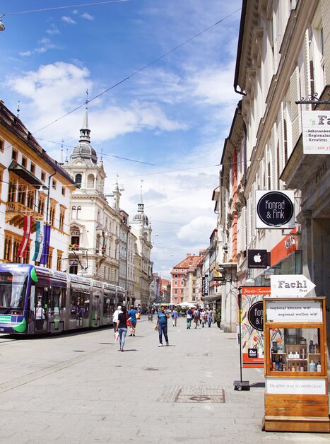 Blick auf eine belebte Straße in Graz mit Geschäften und Straßenbahn. | © sFachl