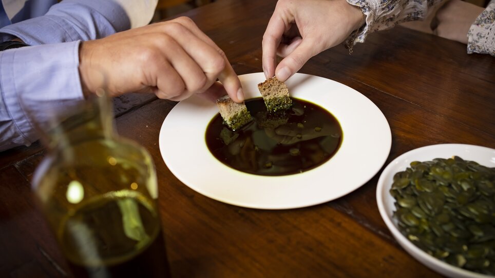 Two people dip bread into a white plate with oil made from Styrian pumpkin seeds. Next to them is a plate of pumpkin seeds and a glass bottle of pumpkin seed oil. | © Graz Tourismus - Werner Krug