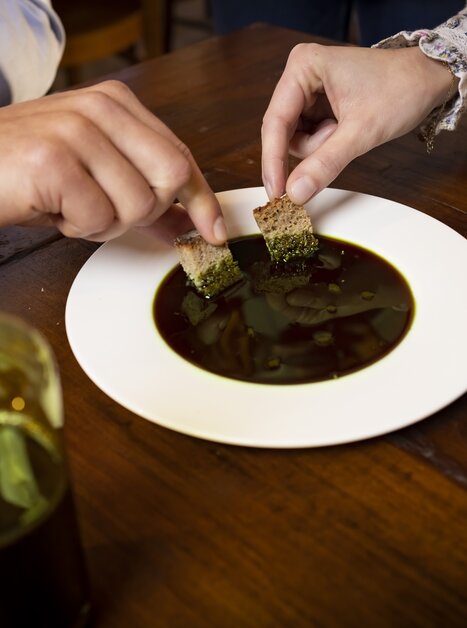 Zwei Personen tauchen Brot in einen weißen Teller mit steirischem Kernöl ein. Daneben steht ein Teller mit Kürbiskernen und eine Glasflasche mit Kernöl. | © Graz Tourismus - Werner Krug