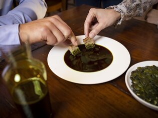 Zwei Personen tauchen Brot in einen weißen Teller mit steirischem Kernöl ein. Daneben steht ein Teller mit Kürbiskernen und eine Glasflasche mit Kernöl. | © Graz Tourismus - Werner Krug