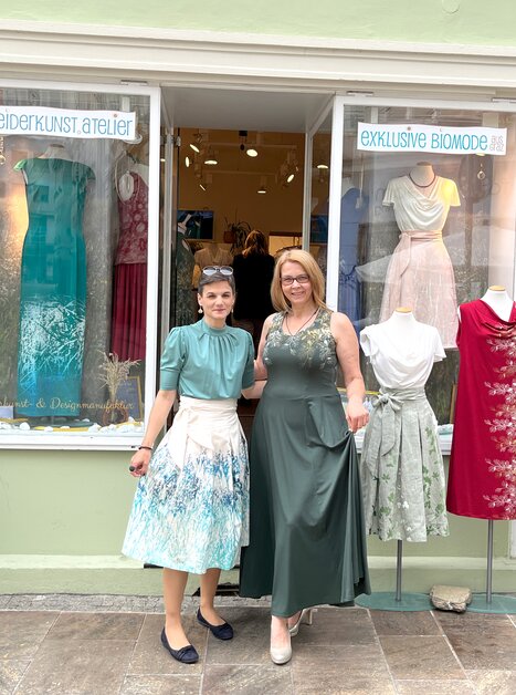 Two women in front of a dress atelier in Graz, surrounded by dresses and plants. | © Peaces