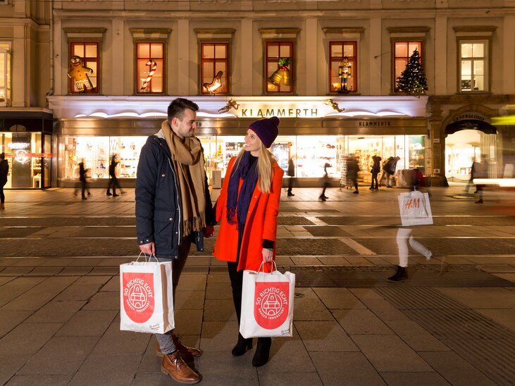 Ein Paar mit Einkaufstaschen vor festlich beleuchteten Geschäften in Graz. | © Harry Schiffer
